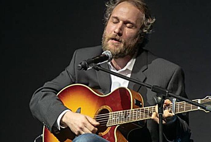 Craig Cardiff sitting on a stool, playing guitar, with a microphone in front.