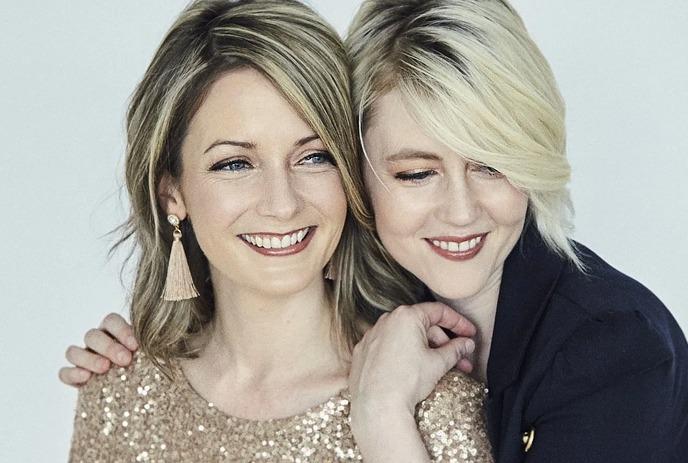 Amanda Walther and Sheila Carabine of the band Dala smiling and hugging each other, in front of a light blue background.