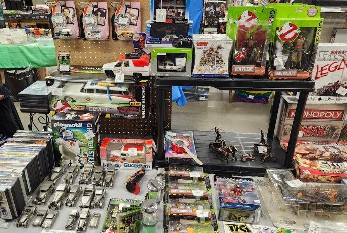 Vintage toy display with Ghostbusters, Monopoly, Playmobil, and die-cast cars on a store table.