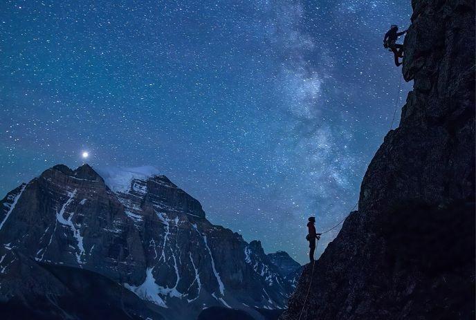 Silhouetted climbers scale a cliff under the Milky Way, with snowy mountains in the background.