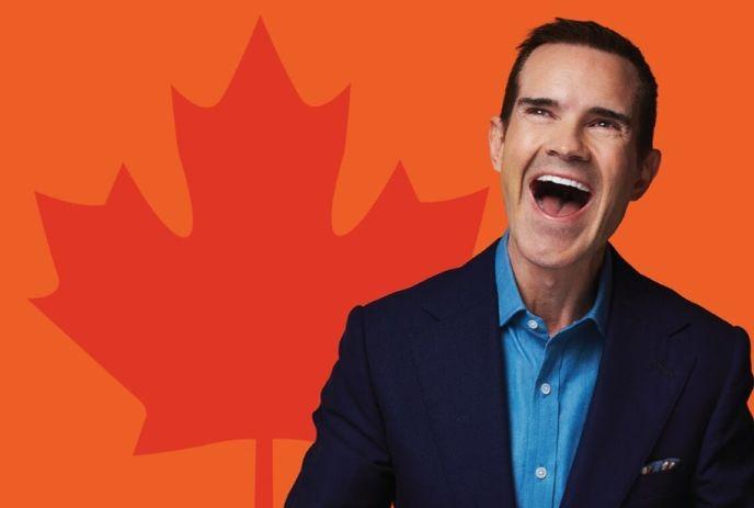 Jimmy Carr wearing a black coat and blue shit, smiling, in front of an orange maple leaf background.