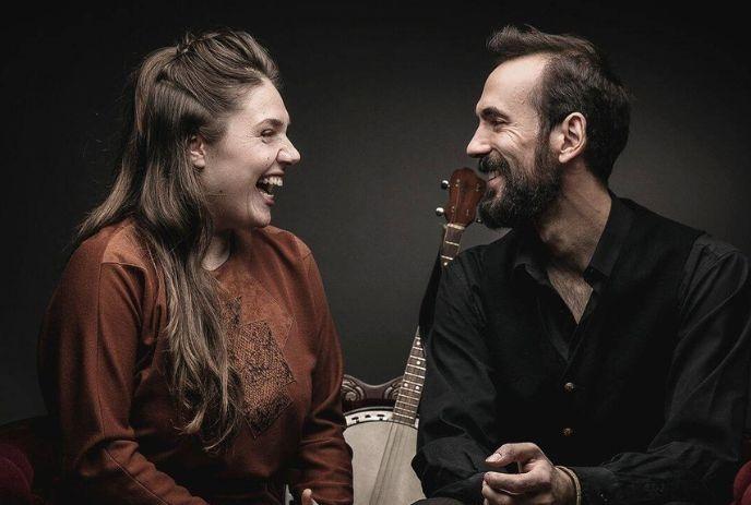 Alex Millaire and Kaitlin Milroy looking at each other smiling, with a banjo behind them, in front of a grey background.