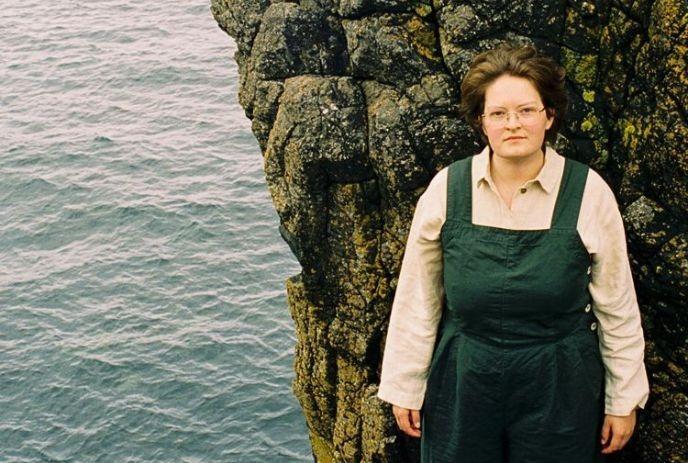 Iona Lane wearing a green dress, standing on a rocky cliff, with water below.