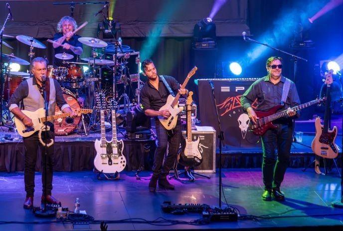 A four-piece rock band performs energetically on stage under colorful lights.