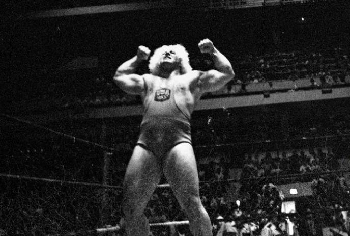 Athlete poses with flexed arms in dark setting, lights highlighting in a wrestling ring.