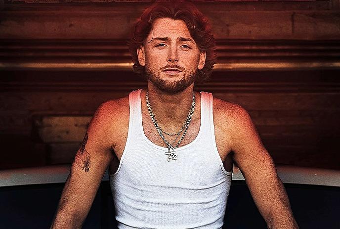 Bailey Zimmerman sits on a seat posing for the camera, wearing a white tank top.