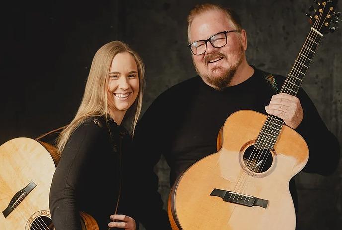 Don Ross & Julie Malia smiling and posing for the camera, while Don holds an acoustic guitar.