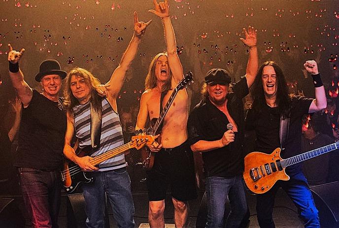 A rock band of five members stands onstage, posing energetically, with instruments, as the crowd cheers.