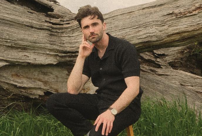 John Muirhead sitting on a stool outside in front of a large tree trunk on a cloudy day.