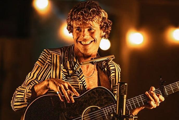 Kim Churchill performing on stage with a harmonica and guitar, in front of a light string.