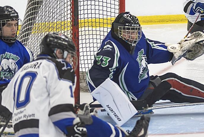 Sled hockey goalie in blue jersey defends net as white jersey player approaches, puck and rink lines visible.