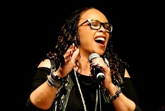 Woman artist in black outfit passionately sings on stage, holding a mic, with braided hair and bracelets, dark backdrop.
