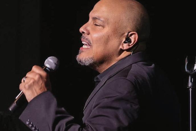 Lead singer in dark suit performs passionately on stage, flanked by two backup vocalists under spotlight.