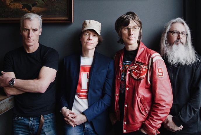 Four band members of Sloan standing side by side, dressed in casual and stylish clothing, with a painting in the background.