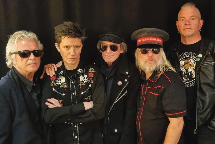 Five artists in dark clothes and shades pose with punk-rock flair; bold jackets, hats, and mohawk styles.