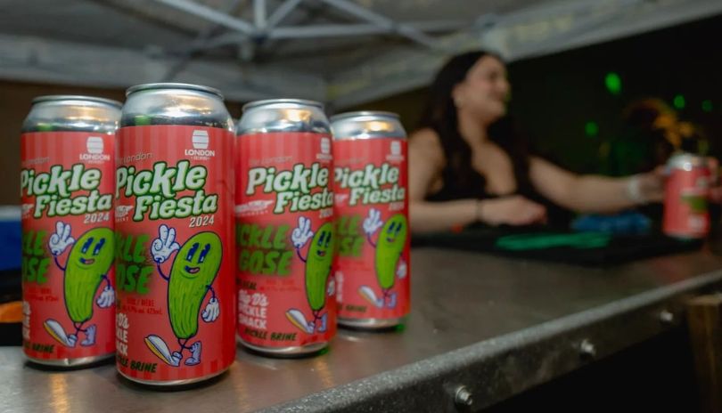 Cans with pink lables saying Pickle Fiesta 2024 under a dark brown atmosphere