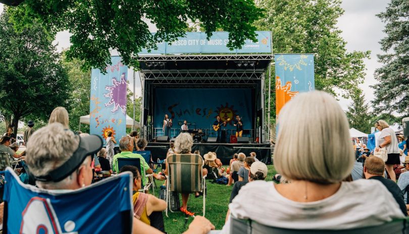 People watch a live band perform on a stage in a park under trees, with a colorful “Sunfest” backdrop and banner.