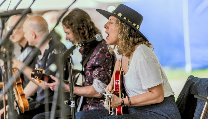 Musicians perform outdoors, playing guitars and singing, with microphones in front, under a large tent.