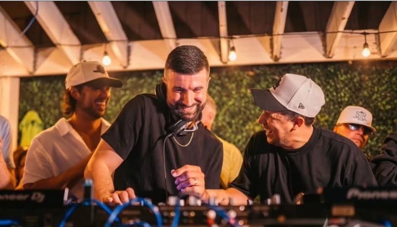 DJ at Pioneer booth with crowd, cables, green wall, NEC logo, party lights—lively event scene.