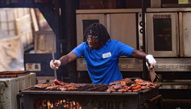 A person in blue shirt and glasses, grilling fresh ribs under a green background.