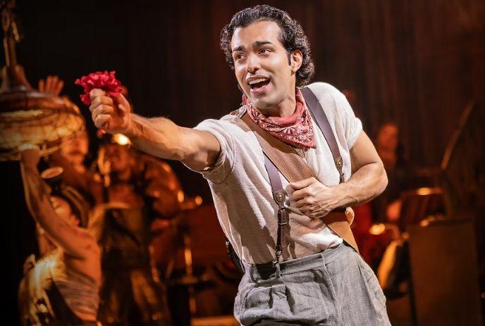 A person in white shirt, gray pants, and red bandana holds a red flower forward, with performers in the background.