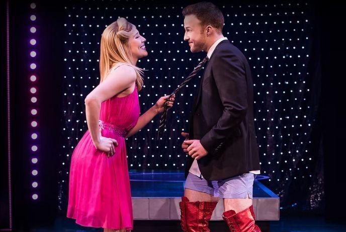 A lady in a pink dress playfully pulls another person's tie. The second person wears a black jacket, white shirt, red boots.