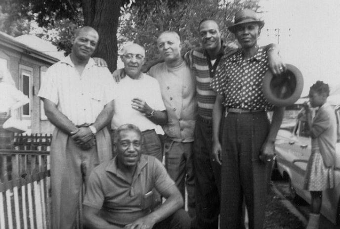 Six men posing outdoors near a tree, with a girl standing by a car in the background.
