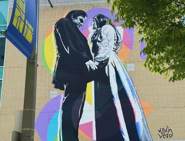 Two figures hold hands in front of vivid shapes: a mural of connection signed by Kevin Ledo on a building wall.