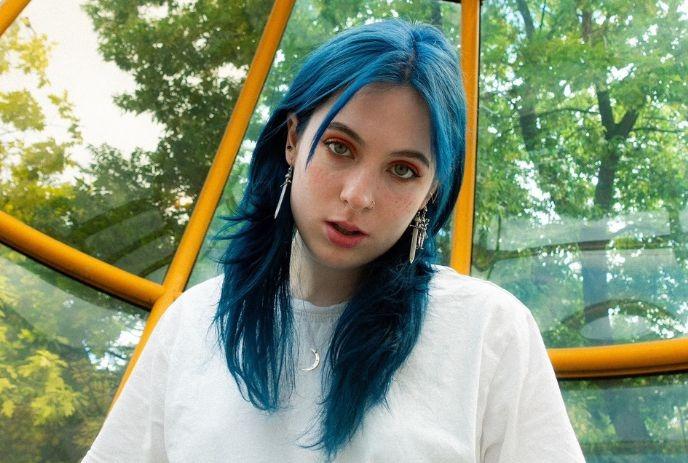 Person with vibrant blue hair stands in front of yellow-framed glass structure, trees visible through background panels.
