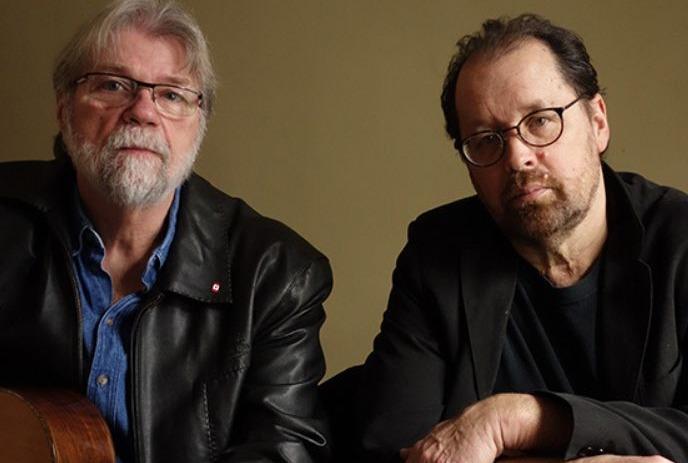 Two artists sit side by side in dark jackets and glasses under a brown background.