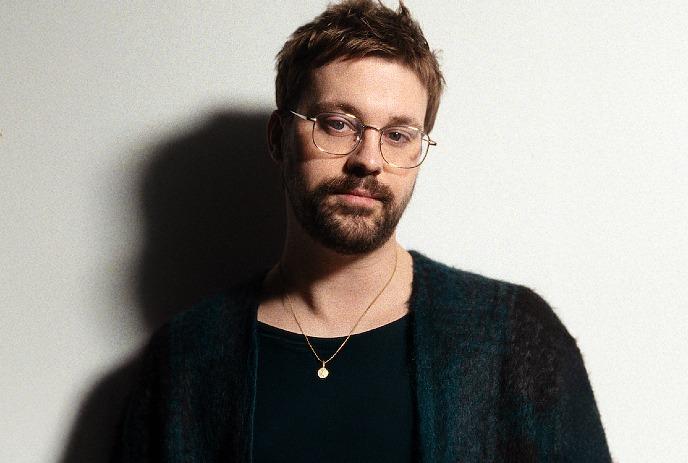 Derik John Baker wearing a sweater and glasses, standing in front of a white background.