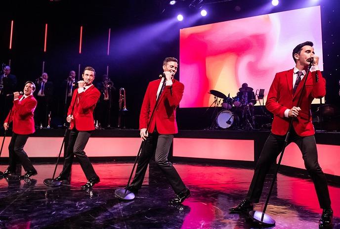 A group of male performers in red suits sing energetically on a vibrant stage.