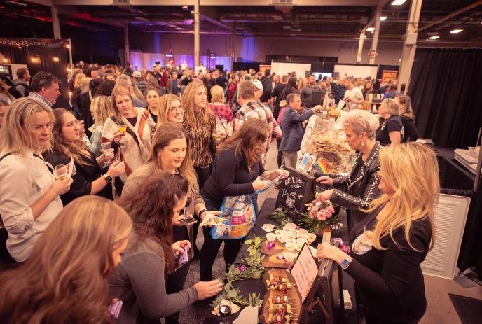 A bustling crowd of attendees samples food and drinks at a vibrant indoor vendor booth in a large convention hall.