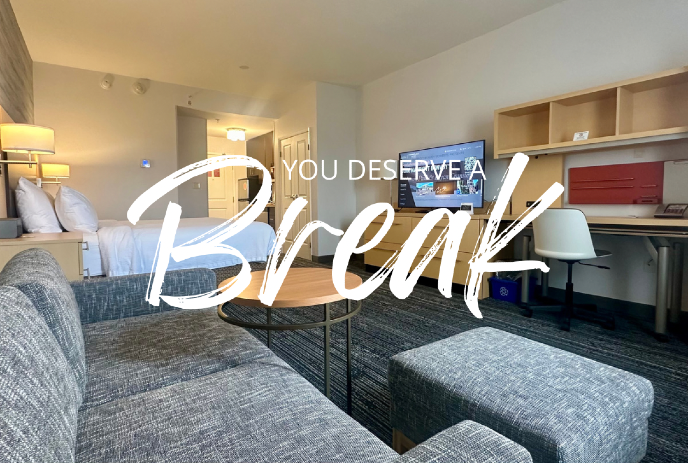 Modern hotel room with bed, sofa, desk, and TV; text overlay reads 'You Deserve a Break'.
