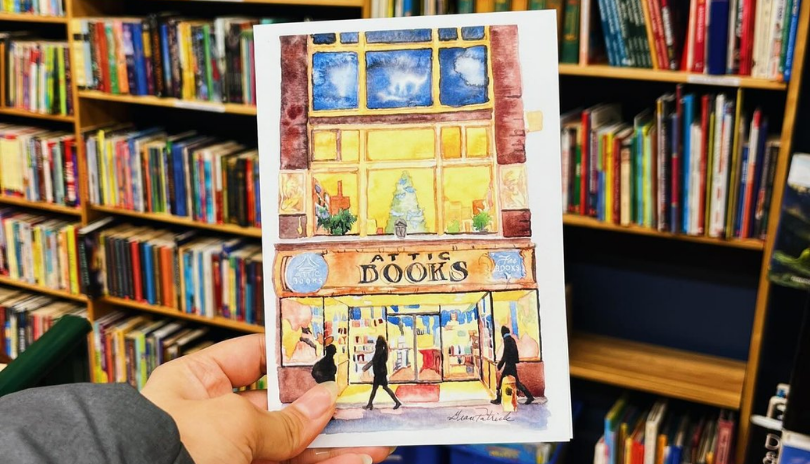 Hand holds an Attic Books illustrated postcard in front of a bookshelf