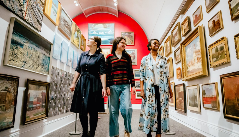 Three adults walk down a hall of paintings in Museum London.