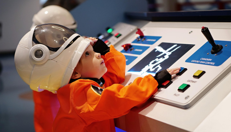 Child in an astronaut outfit stands at a panel of buttons at the London Children's Museum.