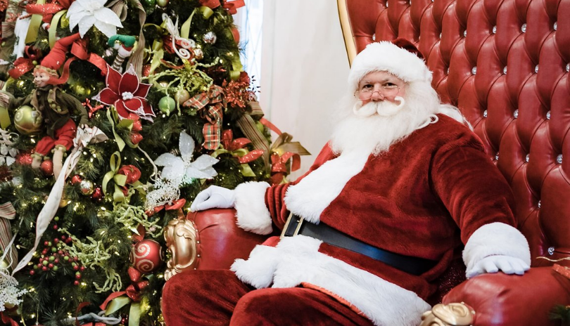 Santa sits on a comfortable red chair next to a beautifully decorated tree.