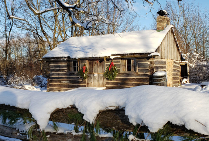Snowy log cabin with wreaths, chimney, and fence; sunlight casts shadows on serene winter landscape.